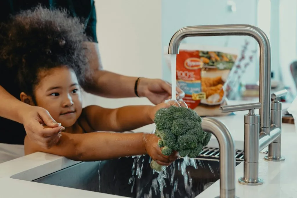 a young girl washing brocolli under a tap